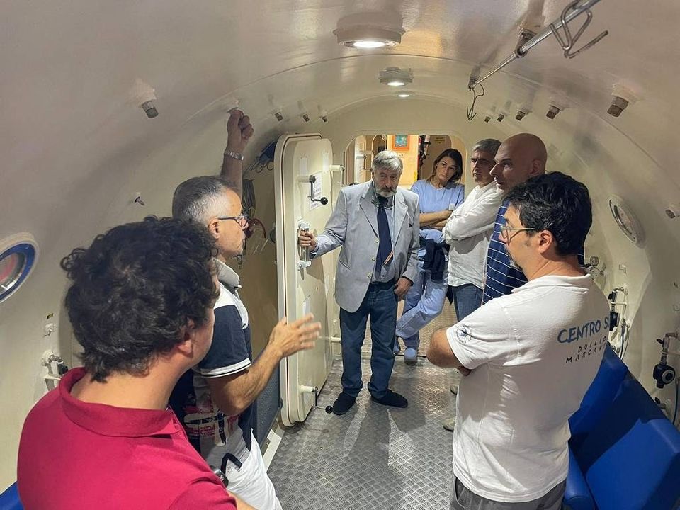 Un gruppo di persone in piedi all'interno di una camera iperbarica. Un uomo al centro, vestito con giacca chiara, sta parlando mentre gli altri ascoltano.